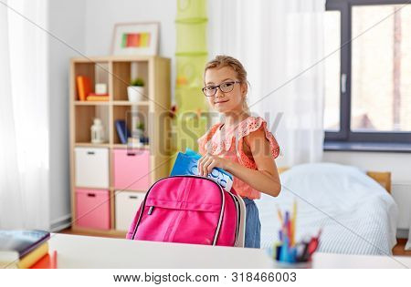 children, education and learning concept - happy student girl in glasses packing school bag at home