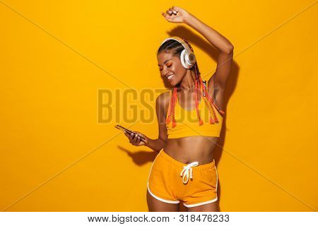 Image of optimistic african american woman singing while listening to music with smartphone and headphones isolated over yellow background