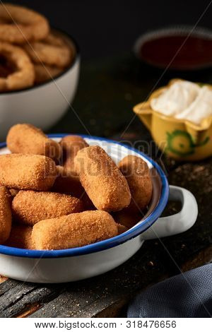 some spanish croquettes in a white and blue enamel plate on a table and some calamares a la romana, fried battered squid rings typical of spain, in the background, next to a mortar with aioli sauce