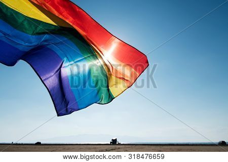 closeup of a rainbow flag waving on the blue sky, moved by the wind, with the sun in the background, on the beach