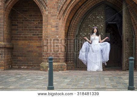 Full Length Portrait Of Caucasian Bride With Crown And Necklace Posing In Church Arch Outdoos.horizo