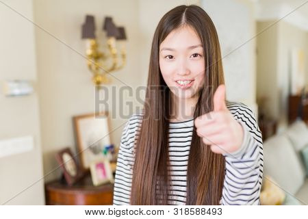 Beautiful Asian woman wearing stripes sweater over living room background doing happy thumbs up gesture with hand. Approving expression looking at the camera showing success.