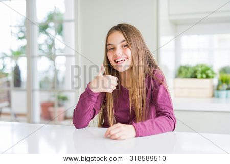 Beautiful young girl kid on white table doing happy thumbs up gesture with hand. Approving expression looking at the camera showing success.