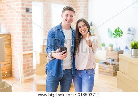 Beautiful young couple moving to a new house using smartphone happy with big smile doing ok sign, thumb up with fingers, excellent sign