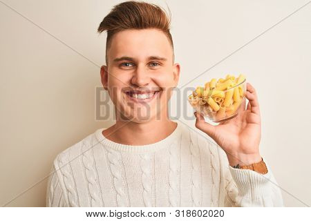 Young handsome man holding bowl with dry pasta standing over isolated white background with a happy face standing and smiling with a confident smile showing teeth