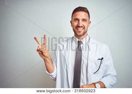 Young handsome doctor man wearing white profressional coat over isolated background smiling with happy face winking at the camera doing victory sign. Number two.