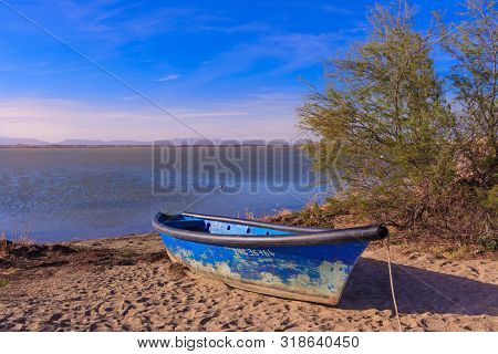 Old Blue Boat On The Beach With Bushes And Calm Water, Blue Sky And Mountains In The Background