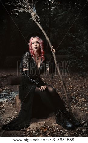 Halloween Theme: Contemporary Young Witch In Black Dress And Hat With Broom Sitting In A Dark Grove