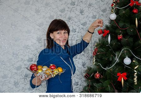 Older Adult Beautiful Brunette Woman In A Casual Style Hanging Balls Decorates A Christmas Tree And 