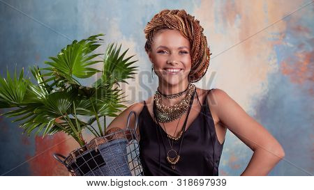 Southern Flavor. A Young Tanned Woman With Afro Braids Smiles Holding A Big Pot With A Tropical Plan