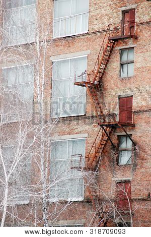 External Fire Escape Staircase On An Old Brick Building Of Factory, Plant. Big Windows, White Birch 