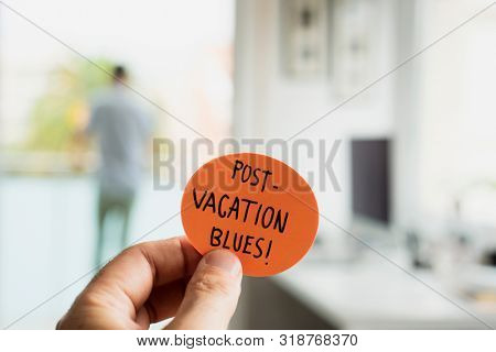 closeup of a man at the office with a note in his hand, with the text post-vacation blues written in it, and a man seen behind standing in the background