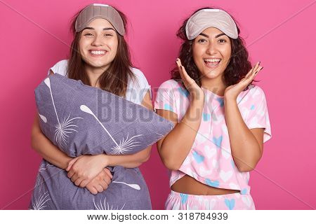 Indoor Shot Of Smiling Women With Sleep Masks On Head, Wearing Pajamas Standing Isolated Over Pink S