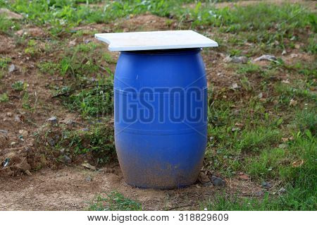 Blue Plastic Water Barrel Covered With Oversize Metal Cover Left In Nature Surrounded With Grass And