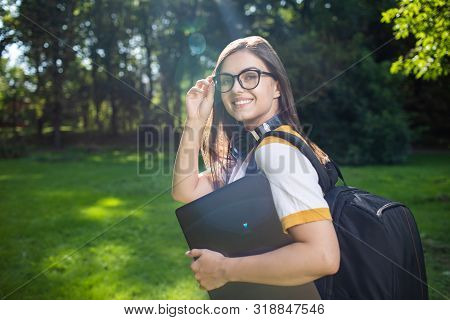 Portrait Of Young Pretty Woman With Laptop And Daypack Adjusting Glasses In Park