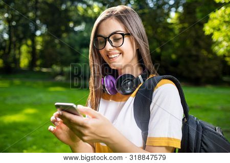 Portrait Of Young Cute Student Girl With Daypack Checking Her Smartphone On The Park Lawn