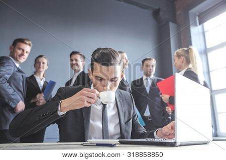 Business Man Working With Laptop And Drinkig Coffee In Office, His Team Of Coworkers On Background