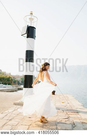 A Gentle Bride In A Dress With An Open Back Is Spinning On The Pier Under The Lighthouse In The Bay 