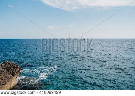 Two White Sailboats Are Sailing On The Open Sea On A Sunny Bright Day. View From The Rocky Shore