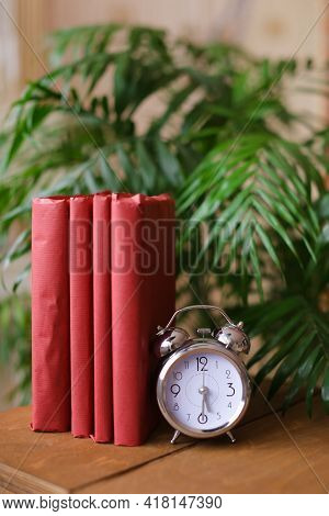 Red Photo Albums Stnding Near Clock And Houseplant On Table.