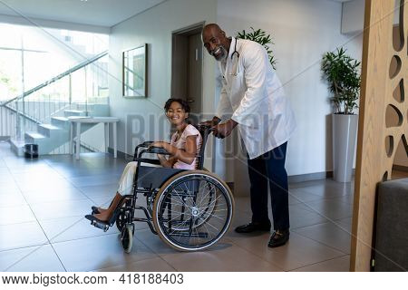 Smiling african american male doctor pushing mixed race girl in wheelchair through hospital corridor. medicine, health and healthcare services.