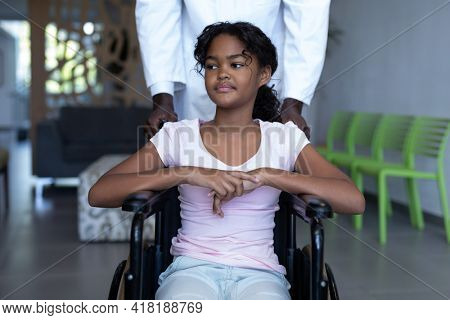 African american male doctor pushing smiling mixed race girl in wheelchair through hospital corridor. medicine, health and healthcare services.