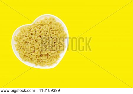 White Heart Shape Plate Filled With Uncooked Pasta Bow Tie On Yellow Background. Butterfly Macaroni,