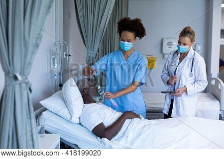Mixed race female doctor wearing face mask putting oxygen mask ventilator on male patient. medicine, health and healthcare services during coronavirus covid 19 pandemic.