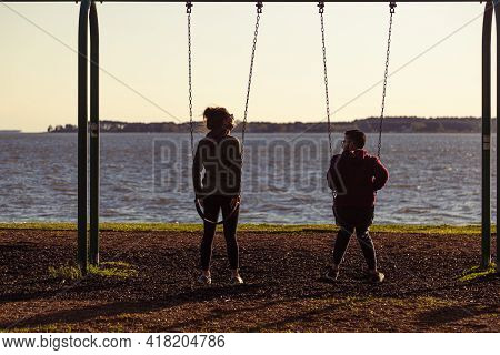 04-16-2021 Oxford, Md, Usa: A Young Couple Is Sitting On Swings At A Playground Overlooking The Wate