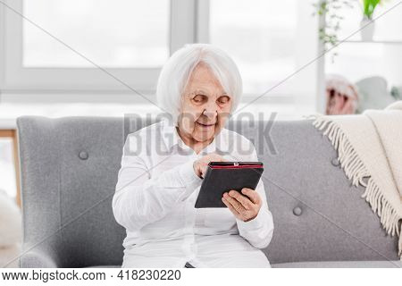 Portrait of elderly gey hair woman sitting on the sofa at home with tablet and looking at screen