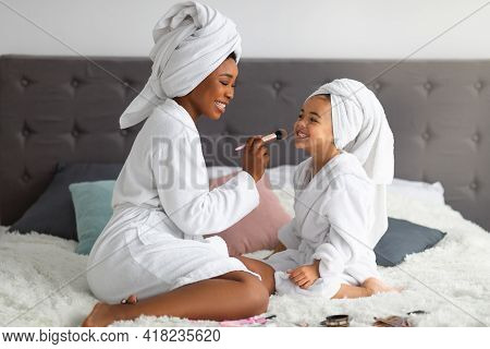 Happy Black Mom And Her Cute Daughter Wearing Bath Robes And Towels, Applying Makeup On Family Spa D