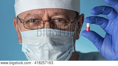 An Elderly Doctor In Medical Clothing: A Cap, Gloves,dressing Gown And Mask, Glasses. He Holds A Med