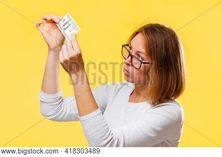 A Pensive Woman Looking At The Dollar Bill Checking For Falsity. Yellow Background. The Concept Of C