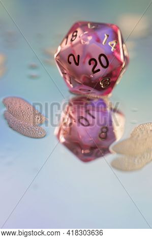 Close Up Of A Pink Transparent D20 Dice On A Wet Reflective Pastel Colered Surface With Foam
