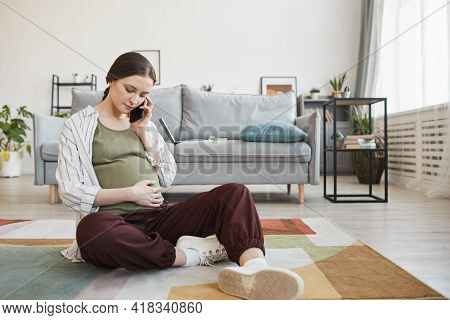 Pregnant Woman Sitting On The Floor And Talking On Mobile Phone In The Living Room