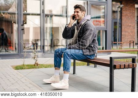 Smiling young white man in casual cothes using mobile phone outdoors in the city, sitting on a bench, talking