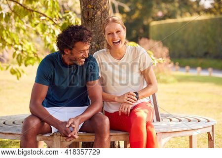 Couple Wearing Fitness Clothing Sitting On Seat Under Tree Checking Activity Monitor On Smartwatch
