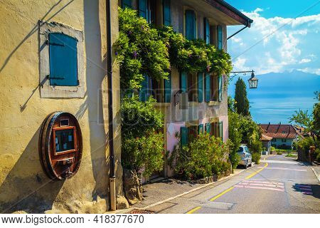 Traditional Swiss House Decorated With Colorful Flowers And Beautiful Street View. Rivaz Village And