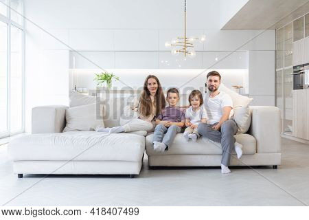 Family Portrait Of Young Parents With Their Two Children At Home, White Modern Desgin Of Living Room