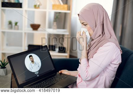 Young Sick Arabian Woman In Hijab, Suffering From Cold And Running Nose, Sits On Couch At Home And U