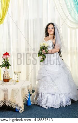 A Happy Jewish Bride Stands In The Hall Before The Chuppa Ceremony At A Table With Flowers With A Bo