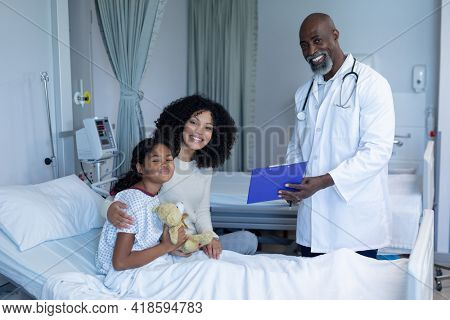 Smiling african american male doctor with mixed race mother and her sick daughter, in hospital bed. medicine, health and healthcare services.