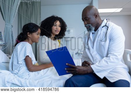 African american male doctor talking to mixed race mother and her sick daughter, in hospital bed. medicine, health and healthcare services.