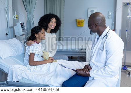 African american male doctor talking to mixed race mother and her sick daughter, in hospital bed. medicine, health and healthcare services.