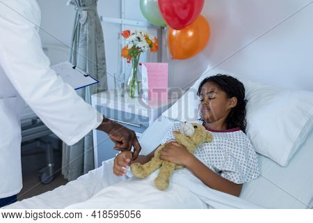 African american male doctor taking pulse of mixed race sick girl asleep in hospital bed. medicine, health and healthcare services.