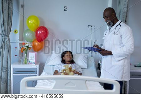 Smiling african american male doctor writing notes by hospital bed of mixed race sick girl patient. medicine, health and healthcare services.