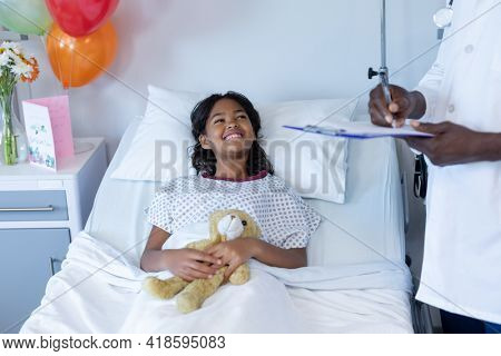 African american male doctor writing at hospital bedside of smiling sick mixed race girl. medicine, health and healthcare services.