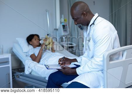 African american male doctor writing notes sitting on hospital bed of mixed race sick girl patient. medicine, health and healthcare services.