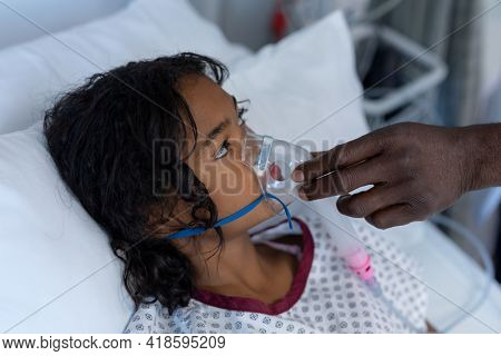 Hand of male doctor putting oxygen mask ventilator on sick mixed race girl in hospital bed. medicine, health and healthcare services during coronavirus covid 19 pandemic.
