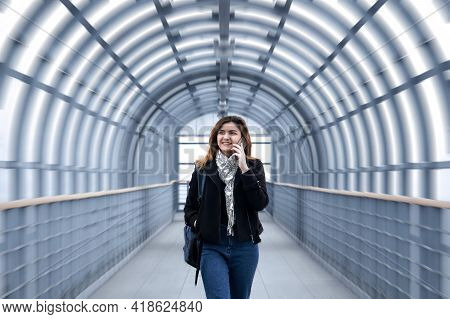 Young Woman Is Walking Rapidly Along A Covered Pedestrian Overpass, Talking On The Phone And Smiling